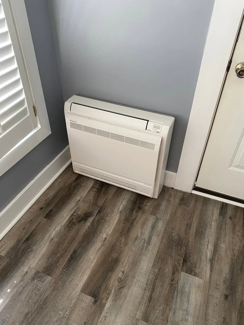 White floor-mounted ductless mini-split installed against a gray wall beside a window.